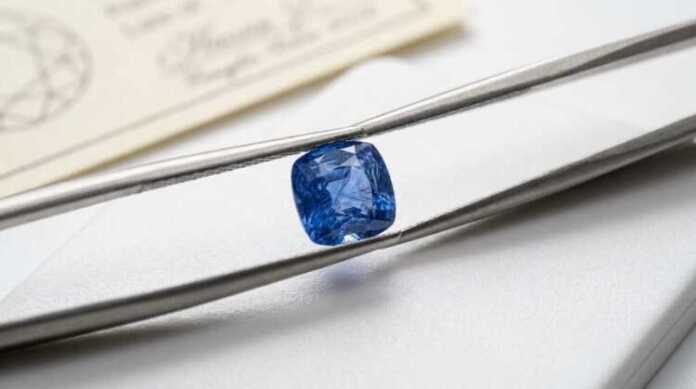 Cushion-cut blue sapphire held in gem tweezers above a grading tray with a gemmological origin report in the background